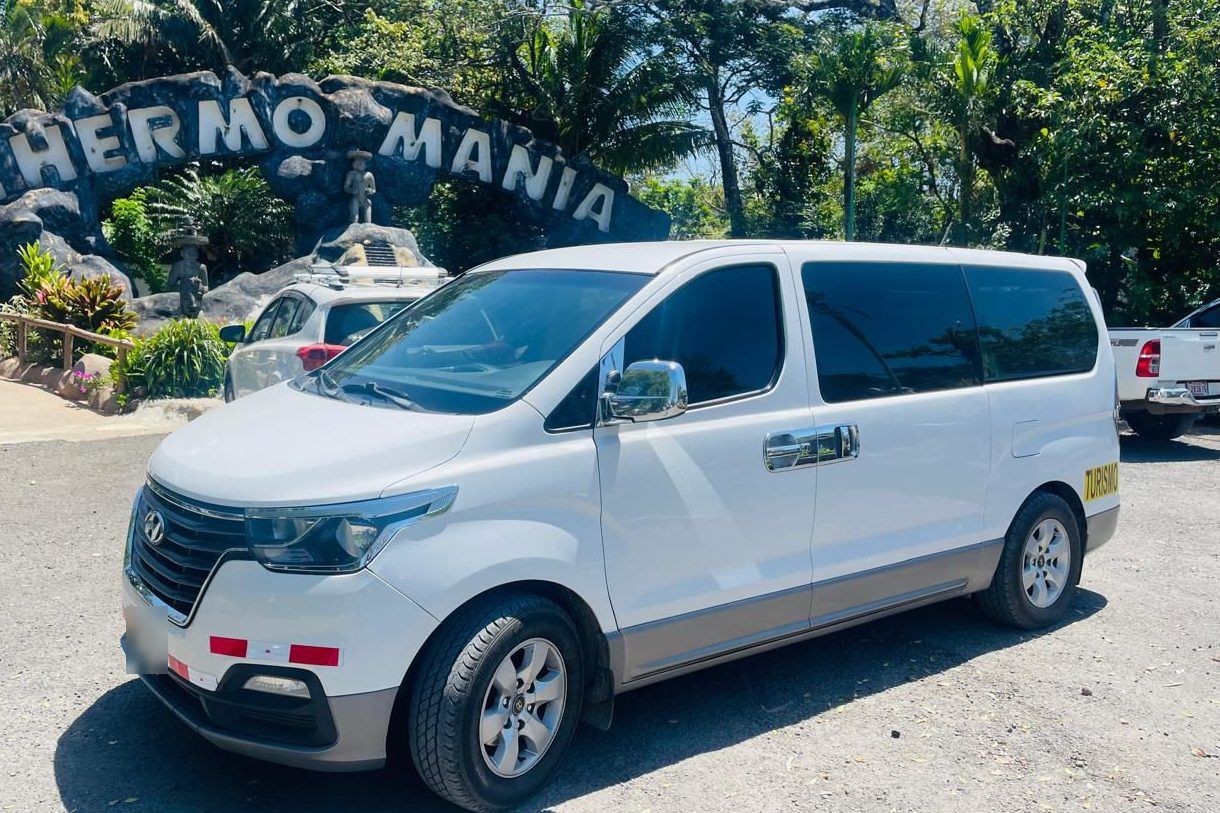 A white Hyundai H-1 van from the fleet parked at the entrance to Thermo Manía hot springs, with the yellow TURISMO tourist-transport licence on its side panel