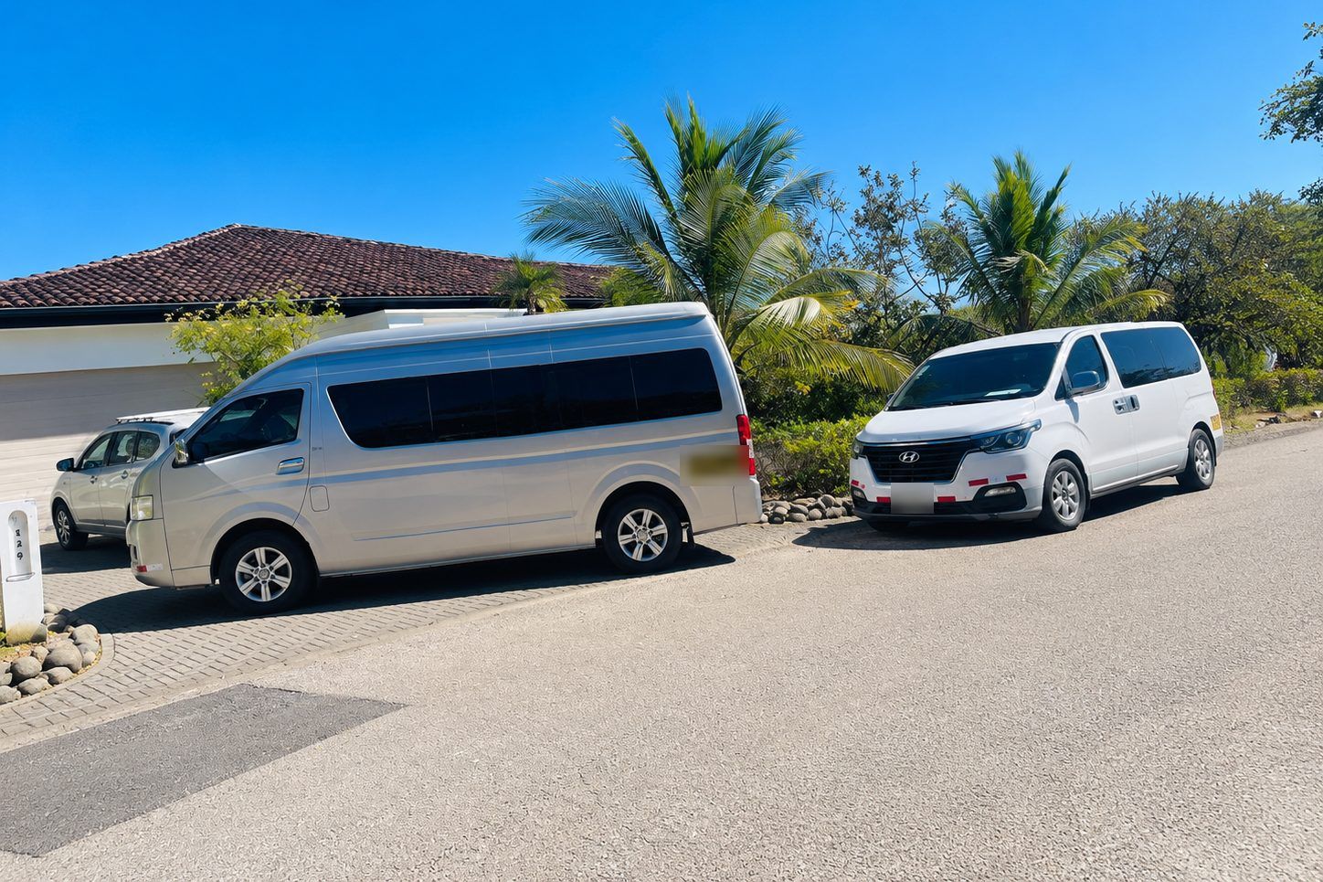 Two Guana Shuttle vans parked on a quiet street in Guanacaste: a silver Toyota Hiace in front and a white Hyundai H-1 behind it